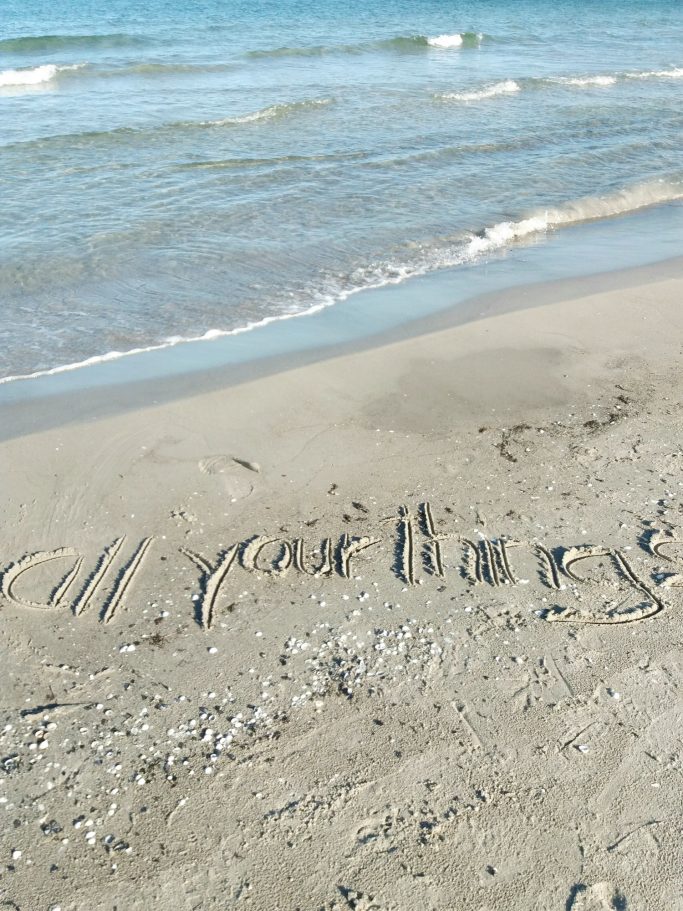 Schriftzug „All you need is love“ im Sand am Strand mit Wellen im Hintergrund.