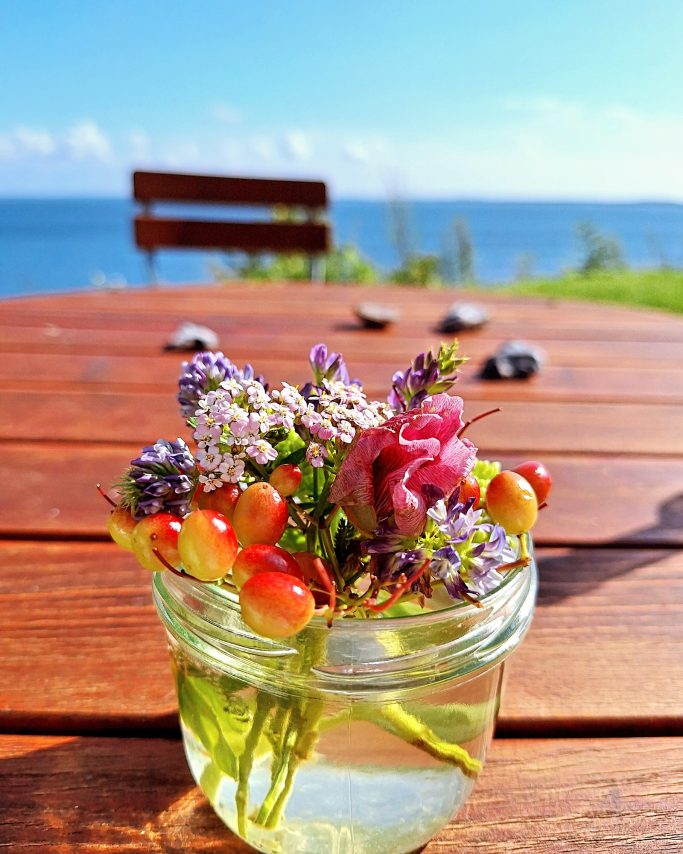 Ein Glas mit bunten Blumen und Beeren auf einem Tisch am Wasser, im Hintergrund ein Holzstuhl.