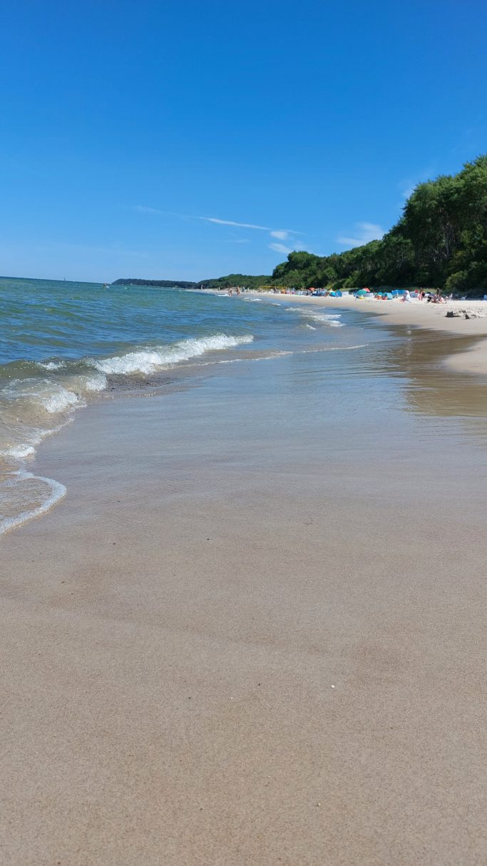 Heller Strand mit sanften Wellen und bewaldeten Hügeln im Hintergrund.