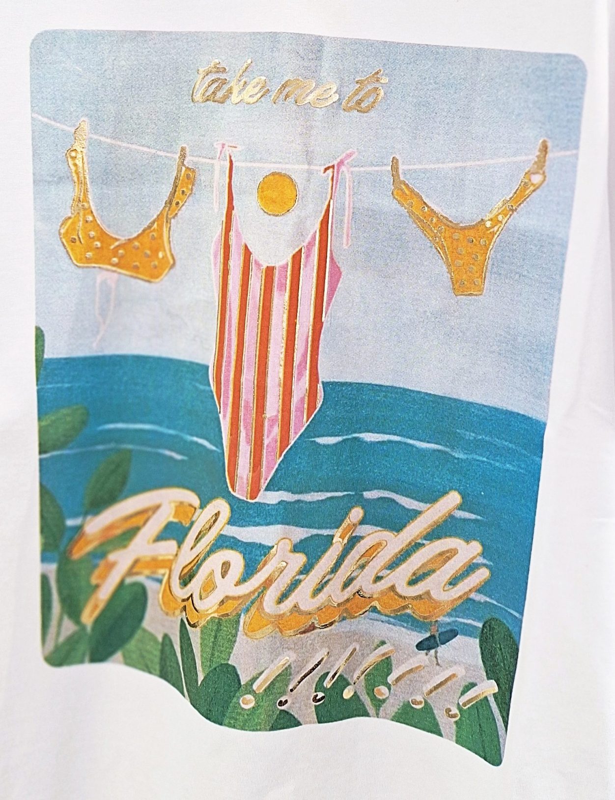 Weißes T-Shirt mit Florida-Aufdruck und Schwimmbikinis vor dem Wasser.