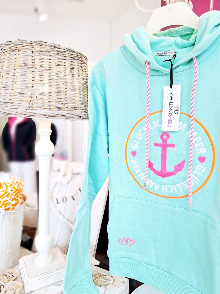 Hellblauer Hoodie mit pinkem Anker-Design und Herzmotiven, neben einer Lampe.