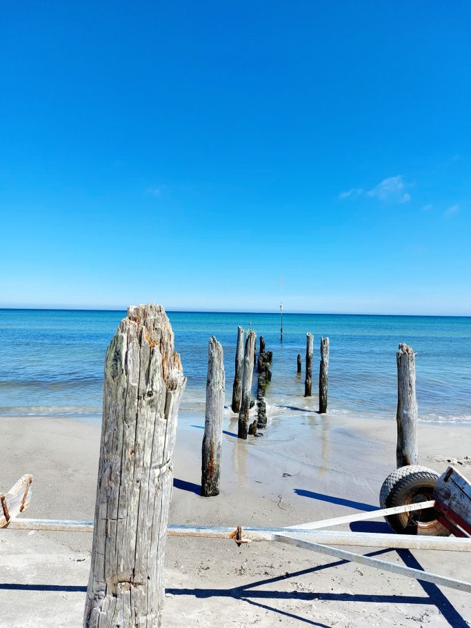 Holzpfähle ragen aus klarem Wasser, blauer Himmel und ruhiger Sandstrand im Hintergrund.