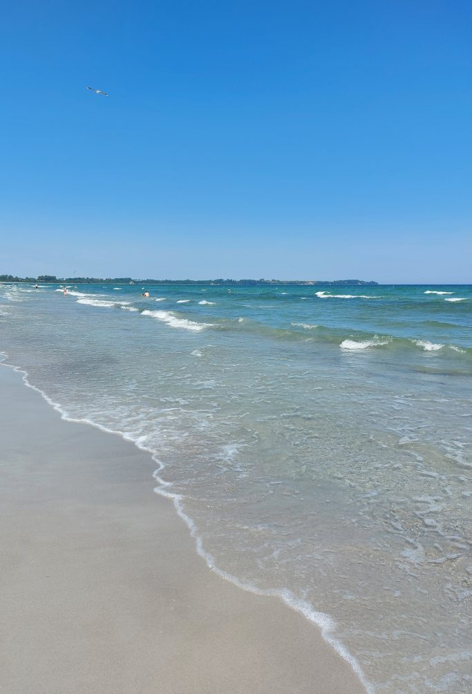 Klares Wasser und sanfte Wellen an einem sonnigen Strand unter blauem Himmel.