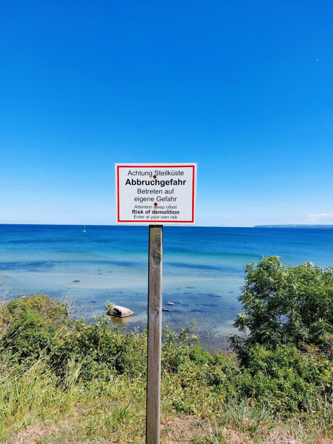 Schild am Ufer, blaugrünes Wasser und klarer Himmel im Hintergrund.