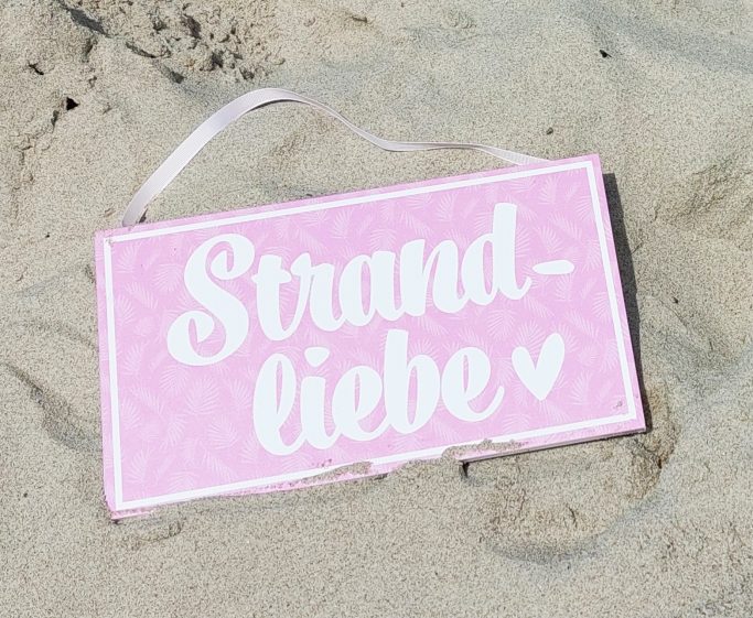 Schild mit der Aufschrift "Strandliebe" in pink auf sandigem Untergrund.