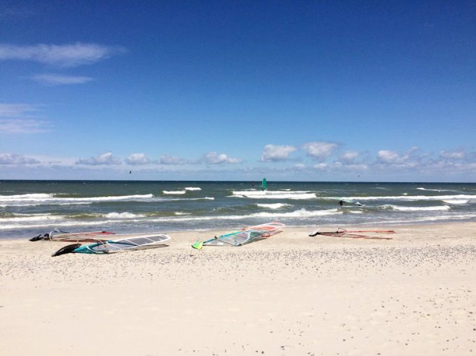 Kitesurfbretter liegen am Strand, Wellen und blaue Himmel im Hintergrund.