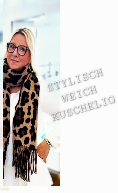 Frau mit blonden Haaren, Brille und Leoparden-Schal, lächelnd in einem hellen Raum.