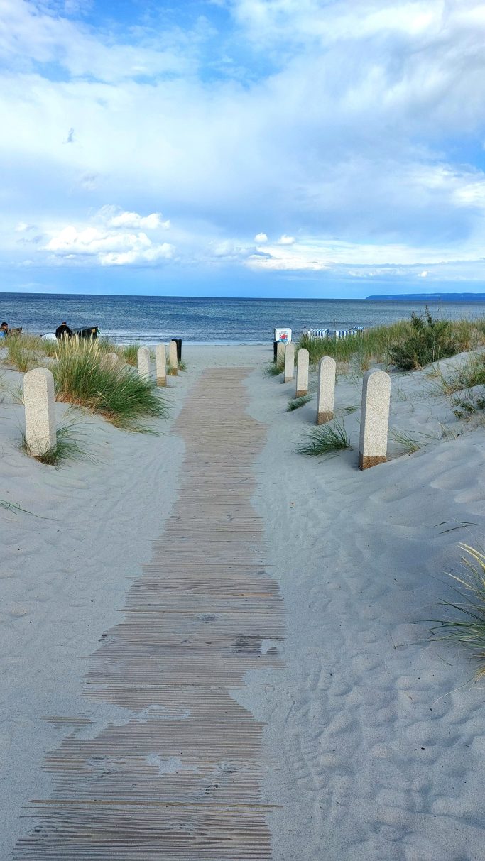 Holzsteg führt durch den Sand zum Strand, umgeben von Gras und leichten Wolken.
