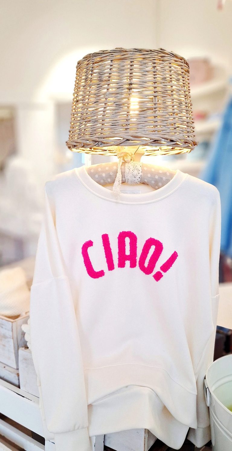 Beige Sweatshirt mit pinkem Schriftzug "CIAO!" hängt neben einer Lampenleuchte.