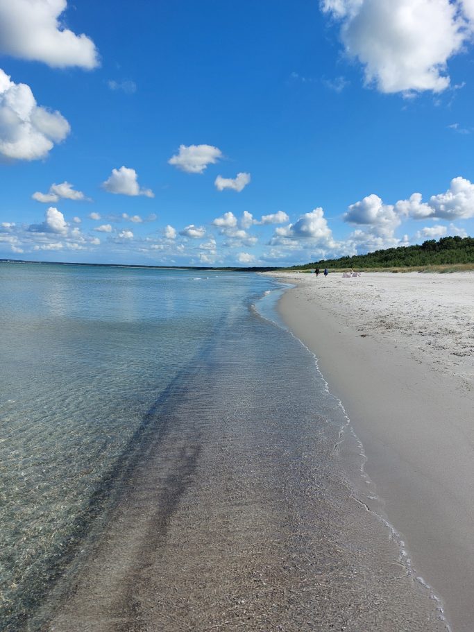 Ruhiger Strand mit klarem Wasser, feinem Sand und bunten Wolken am Himmel.