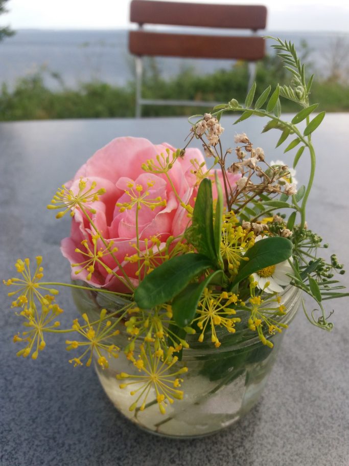 Eine rosa Blume mit gelben und grünen Pflanzen in einem Glasbehälter.
