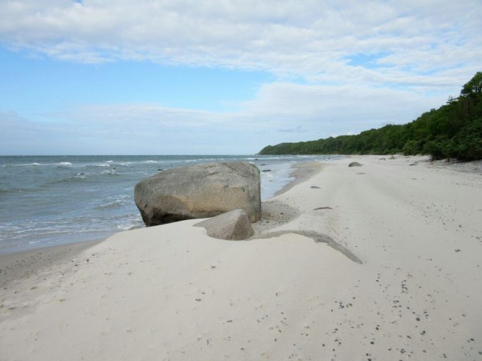 Ein ruhiger Strand mit feinem Sand, einem großen Felsen und sanften Wellen.