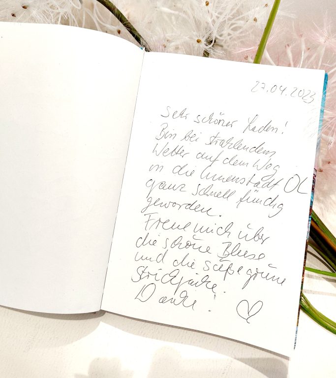 Handschriftliche Notiz in einem offenen Notizbuch mit blühenden Pflanzen im Hintergrund.
